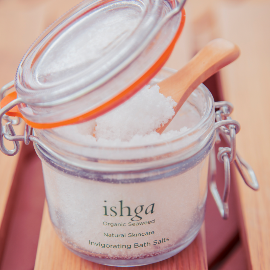 Glass jar of ishga Invigorating Bath Salts with lid open and a small wooden spoon