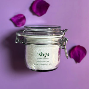 Glass jar of ishga Invigorating Bath Salts with small wooden spoon and sea salt flakes next to it