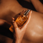 A hand holding a bottle of Ishga over a person's shoulder, with warm, dramatic lighting highlighting the golden liquid and skin tones against a dark background.