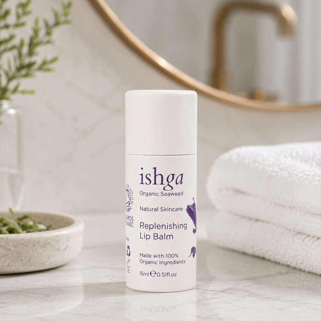 A white bottle of ishga Replenishing Lip Balm sits on a marble bathroom counter with a folded white towel, a small plant, and a round mirror in the background.