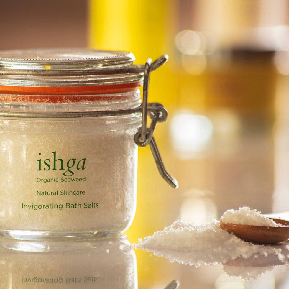 Glass jar of ishga Invigorating Bath Salts with small wooden spoon and sea salt flakes next to it
